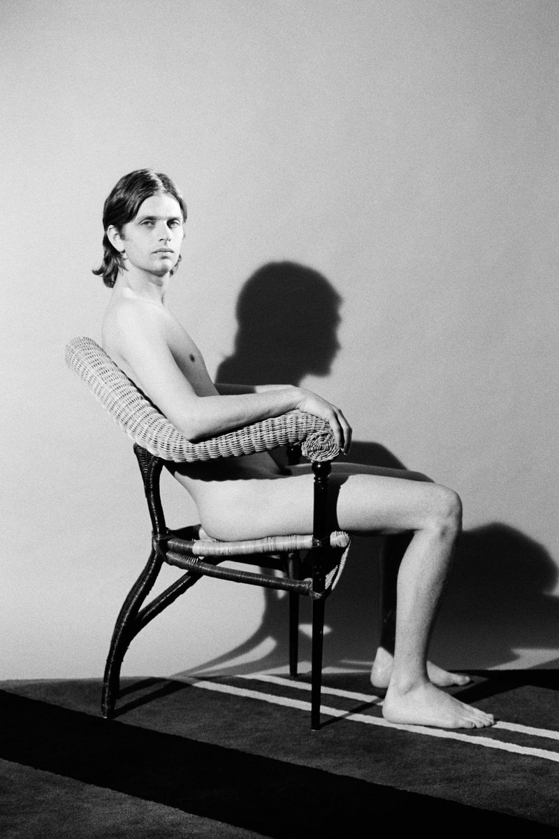 Nude young man seated in profile on a wicker armchair against a plain wall casting a defined shadow.