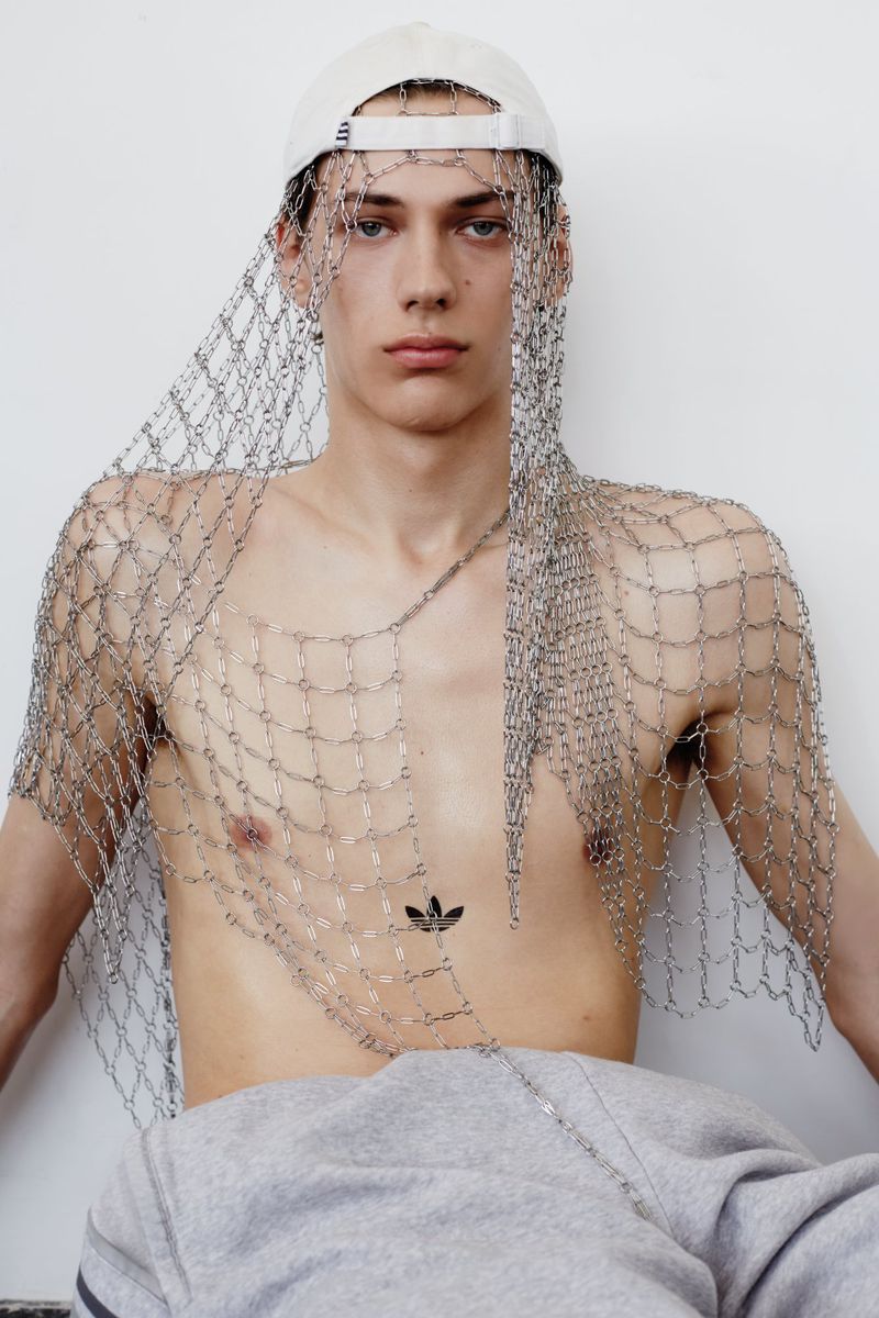 Shirtless male model wearing a metallic chainmail net veil and backward white cap against a plain white background.