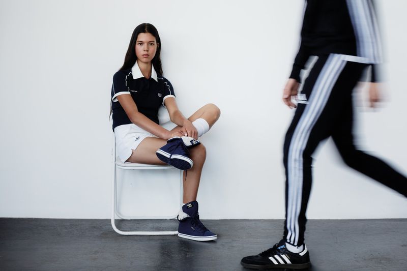 A seated woman in a navy polo shirt and shorts looks forward as a motion-blurred person in track pants walks past against a white wall.