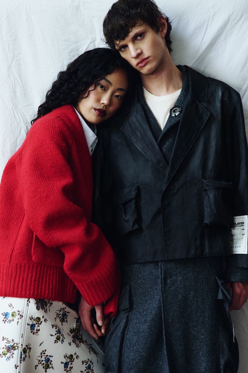 A female model in a bright red knit jacket leaning against a male model wearing a dark structured utility blazer against white draped fabric.