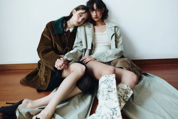 Two models seated on a wood floor leaning against each other wearing oversized trench coats and patterned boots.