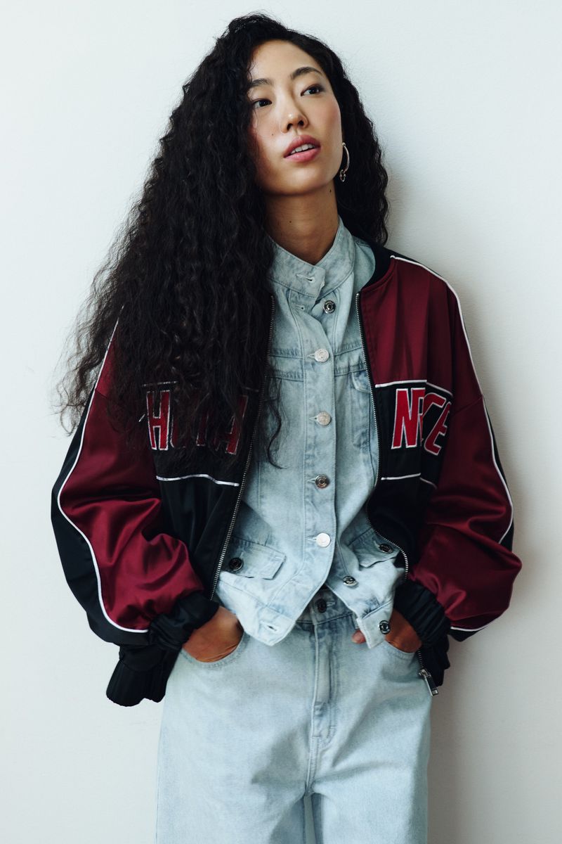 Model Kris Wardak with curly hair posing in a denim ensemble and a burgundy satin bomber jacket against a white wall.