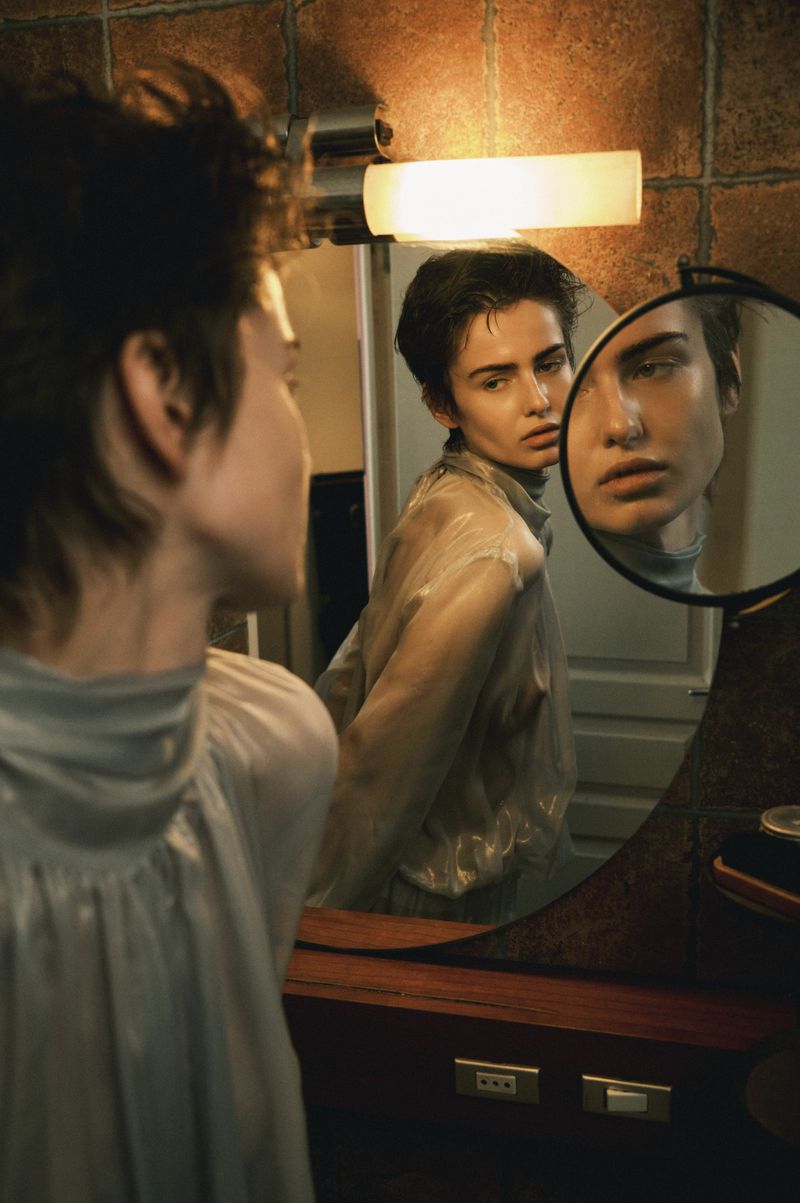 Model with short dark hair in a sheer high-neck top reflected in dual vanity mirrors under warm bathroom lighting.