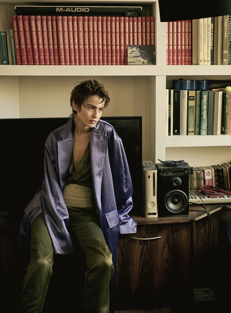 Model Lando van der Schee seated in a purple satin coat and olive trousers in front of a bookshelf.