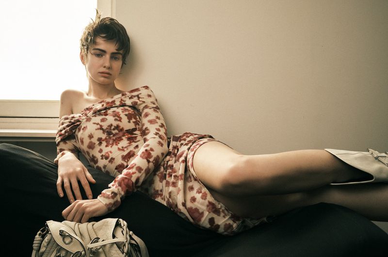 Model Lando van der Schee reclining on a dark sofa in a floral off-the-shoulder dress next to a cream leather handbag.