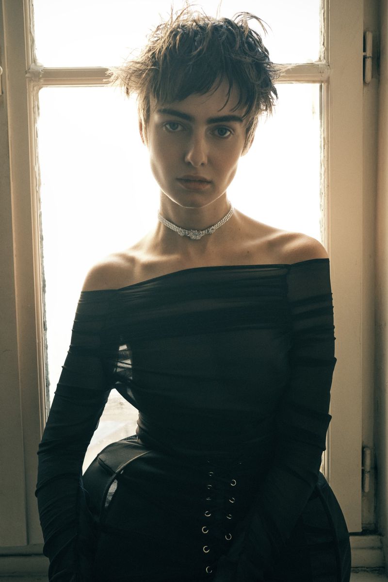 Model Lando van der Schee wearing a sheer off-the-shoulder black top and sparkling choker in front of a bright window.