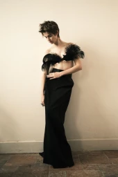 Model Lando van der Schee posing against an off-white wall wearing a black feather bralette and a floor-length black skirt.