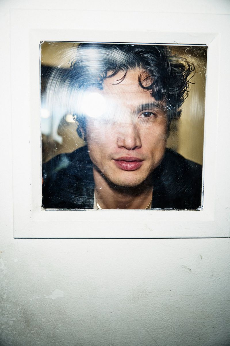 Actor Charles Melton looking directly forward through a square glass window framed in a white textured wall.