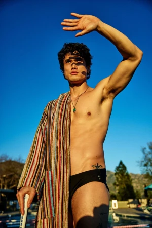 Actor Charles Melton standing poolside wearing a striped bathrobe draped over his shoulder and black swim briefs under a clear blue sky.