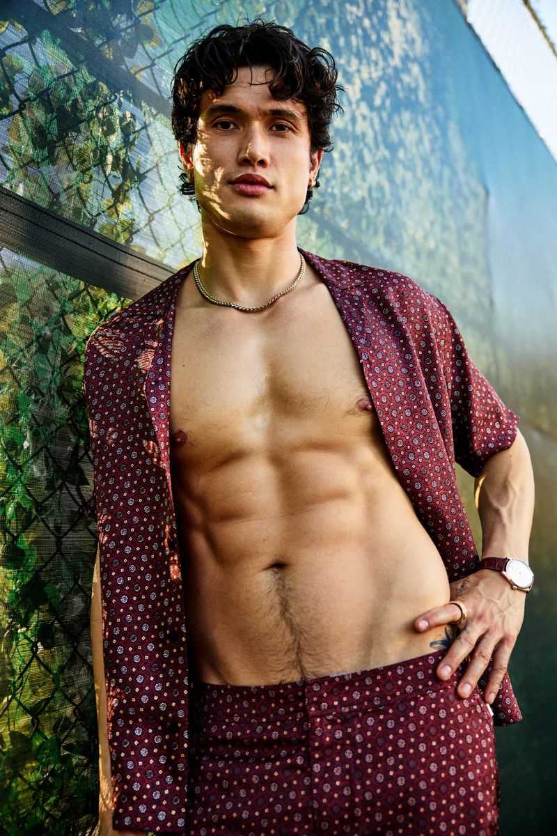 Charles Melton poses shirtless wearing an unbuttoned burgundy patterned shirt and matching pants outdoors against an ivy fence.