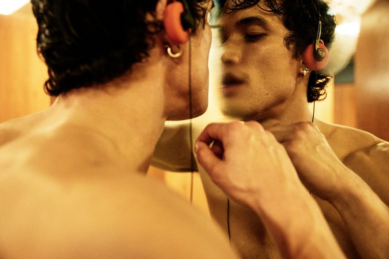 Shirtless Charles Melton wearing retro orange foam headphones and gazing at his reflection in a warm-lit mirror.