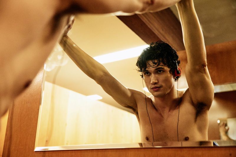 Actor Charles Melton shirtless looking into a mirror with arms raised while wearing wired headphones with red ear pads.