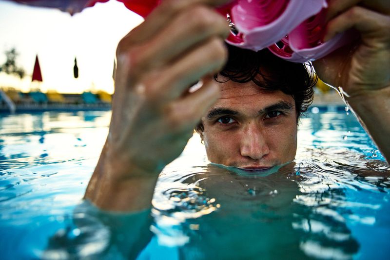 Close-up portrait of actor Charles Melton in a swimming pool holding pink goggles above the water for Men's Health U.S.