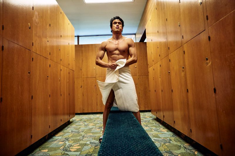 Charles Melton wearing a white bath towel in a wood locker room for Men's Health U.S.