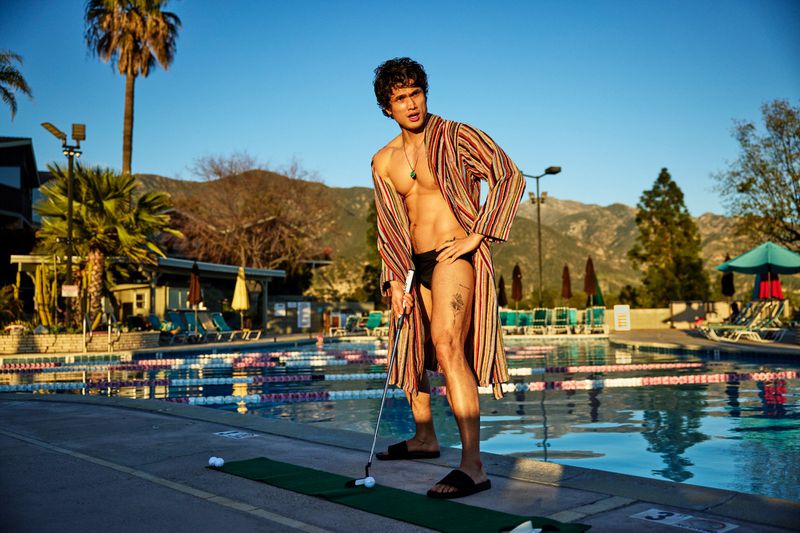 Charles Melton wearing an open striped bathrobe and swim briefs while putting a golf ball by an outdoor swimming pool.