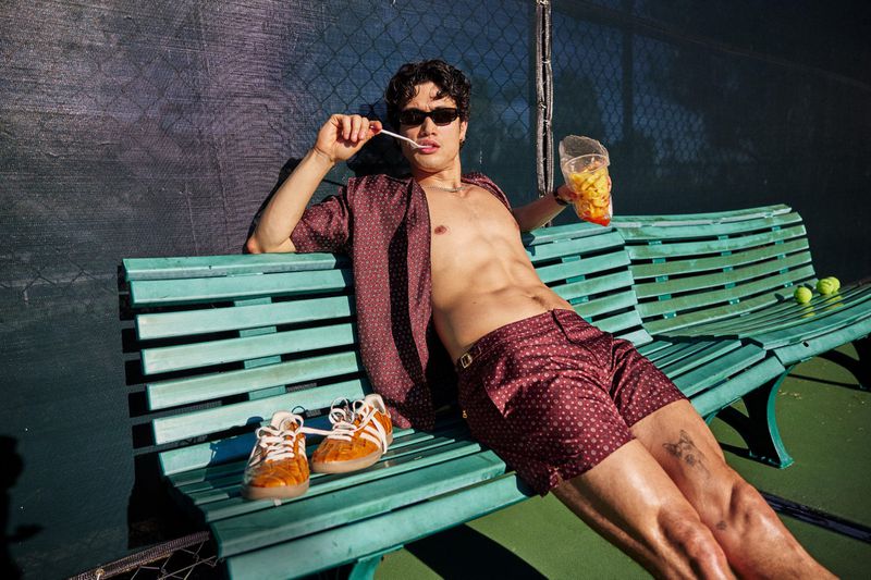 Actor Charles Melton lounging shirtless in burgundy patterned shirt and shorts on a green bench at a tennis court.