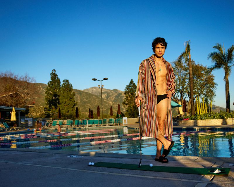 Charles Melton wearing an open striped bathrobe and black slides holding a golf putter by an outdoor swimming pool.