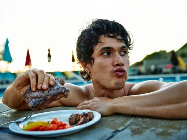 Actor Charles Melton resting at a poolside holding a grilled steak next to a plate of food.