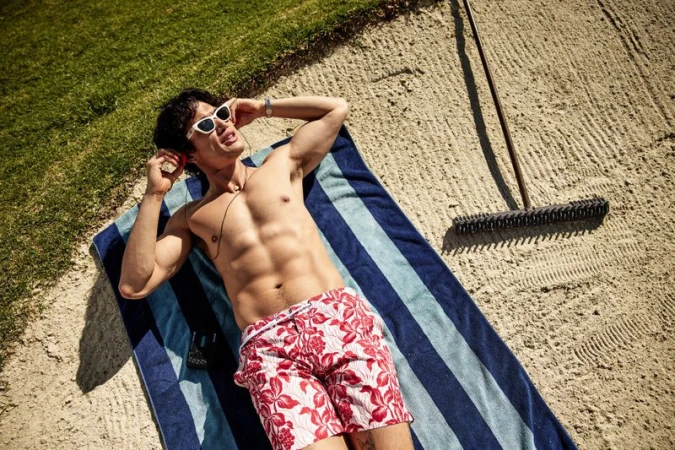 Charles Melton lying shirtless in a sand trap on a blue striped towel wearing red floral swim trunks and sunglasses.