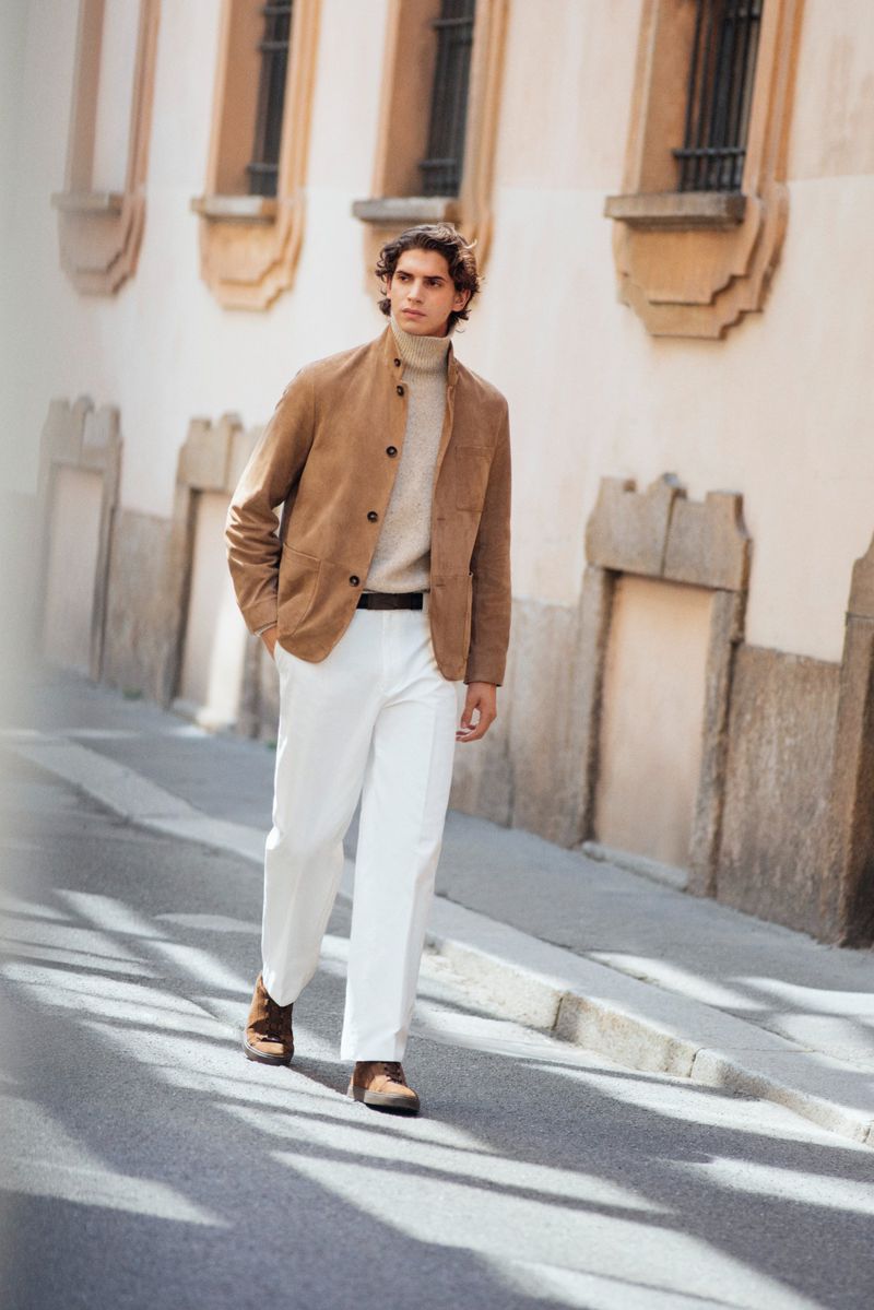 Model Hedi Ben Tekaya walks along a sunlit street wearing a tan suede jacket, cream turtleneck sweater, and white wide-leg trousers.