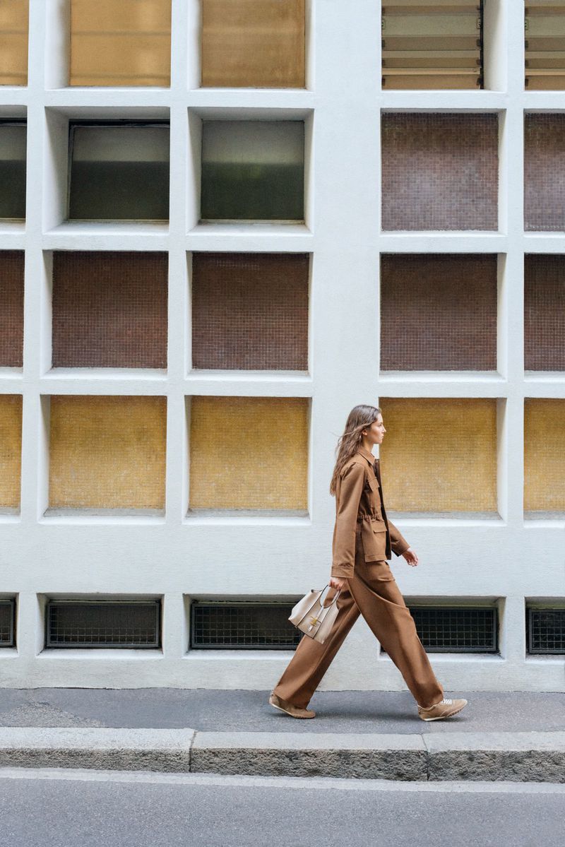 Model in a brown suit walking along a sidewalk holding a beige handbag against a geometric window grid facade.