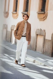 Model Hedi Ben Tekaya walks along a sunlit street wearing a tan suede jacket, cream turtleneck sweater, and white wide-leg trousers.