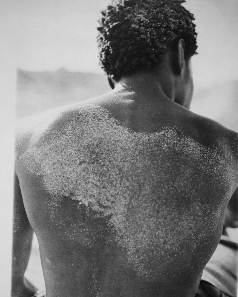 Monochrome, medium shot of a Black adult male from the back, covered in sand at the beach. His head is turned slightly left.