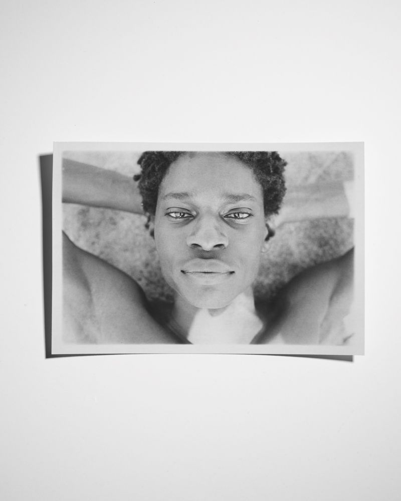 Monochrome portrait of an adult Black woman lying down with her hands behind her head, photographed indoors with soft lighting.