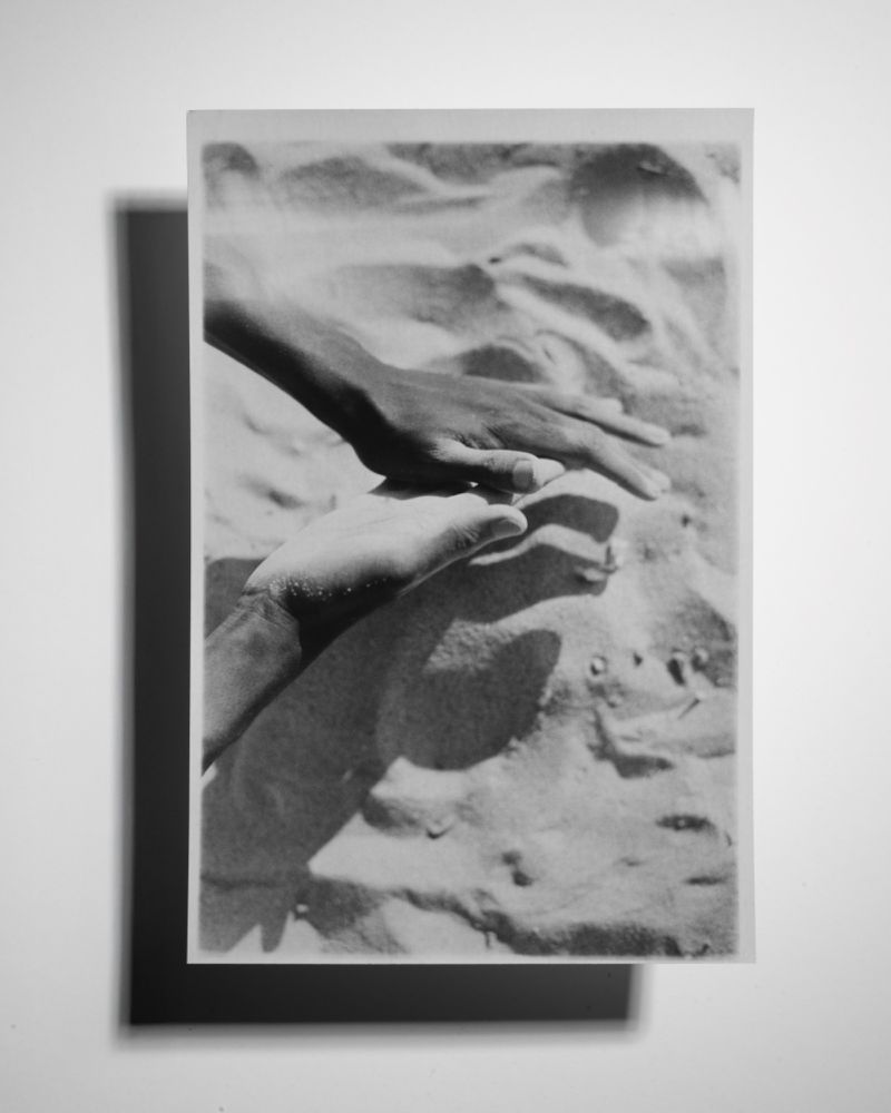 Monochrome photograph of two human hands touching on a sandy surface with shadows, presented as a print on a white background.