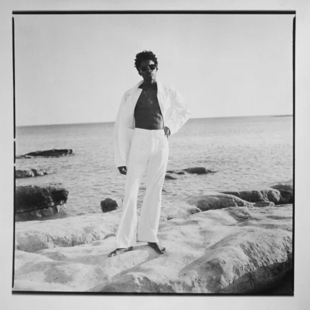 An adult male poses on rocks near a sea. He wears sunglasses, an unbuttoned white jacket and white pants. The sky is bright and clear.