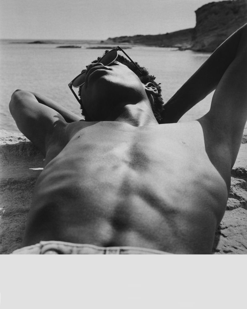 Monochrome close-up of a shirtless Black man wearing dark sunglasses lying back on a sandy beach under direct sunlight.