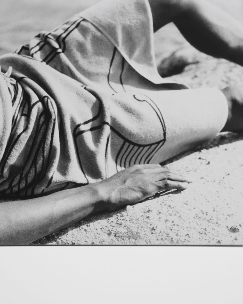 Monochromatic image of a person lying on a blanket with linear patterns, resting on a stone surface under bright, neutral lighting.