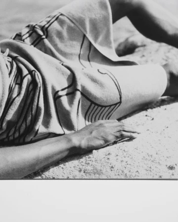 Monochromatic image of a person lying on a blanket with linear patterns, resting on a stone surface under bright, neutral lighting.