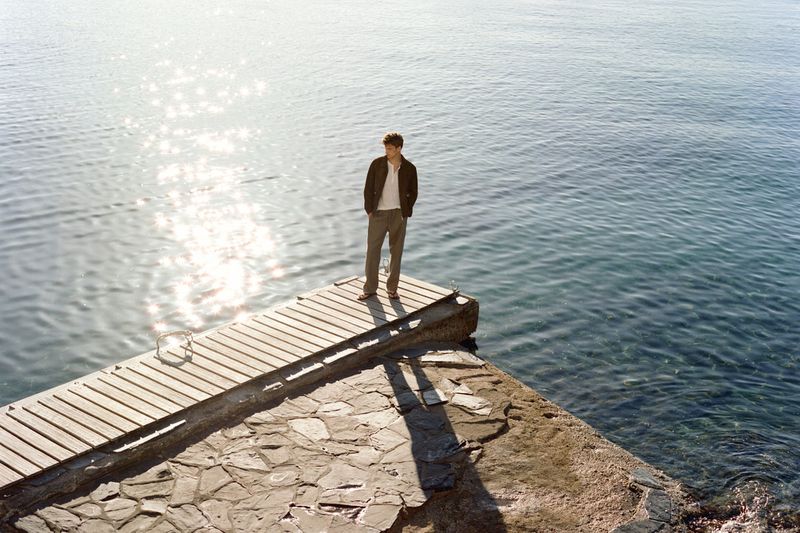 A man in a dark jacket and khaki trousers stands at the edge of a wooden dock overlooking sparkling ocean water.
