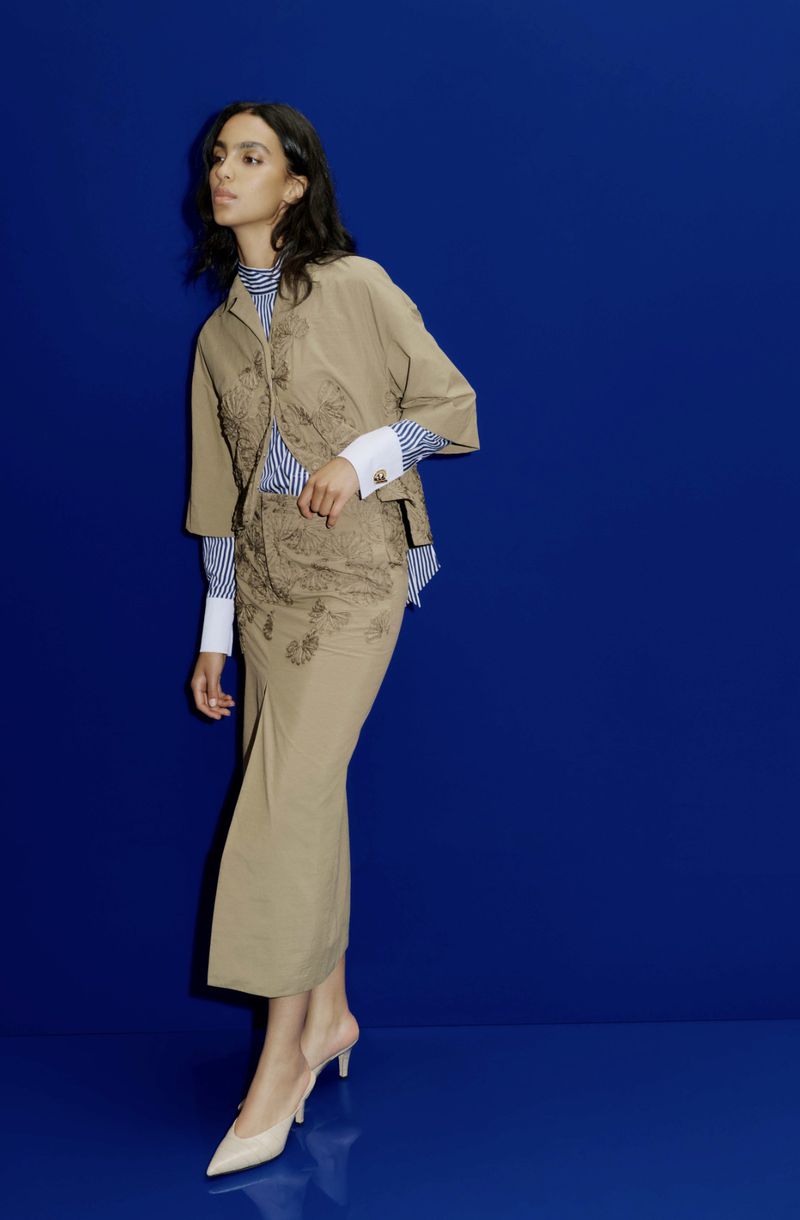 Model Noemi Kassa wearing a tan embroidered jacket and midi skirt set against a vibrant royal blue studio backdrop.
