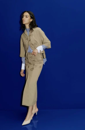 Model Noemi Kassa wearing a tan embroidered jacket and midi skirt set against a vibrant royal blue studio backdrop.