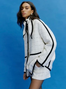 Model Noemi Kassa posing in a white tweed jacket with black piping and matching shorts against a solid blue studio backdrop.