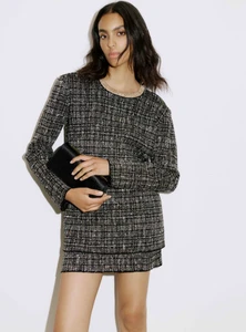 Model Noemi Kassa poses in a long-sleeve tweed mini dress holding a black clutch against a plain white studio backdrop.