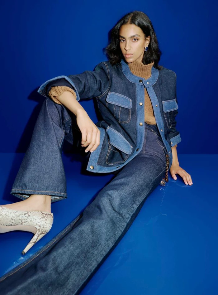 Model Noemi Kassa wearing a paneled denim jacket, wide-leg jeans, and snakeskin heels against a cobalt blue studio backdrop.