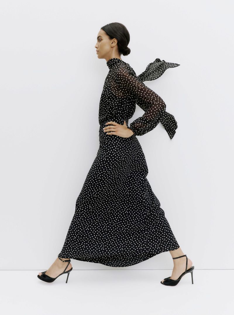 Model Noemi Kassa walking in profile wearing a black polka dot maxi dress and black heels against a white backdrop.