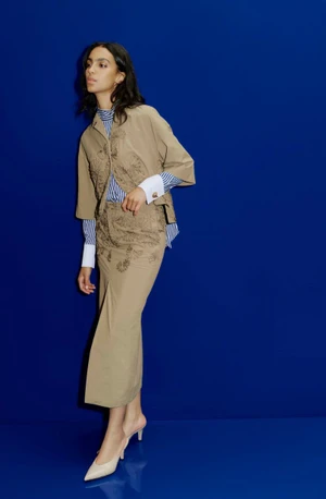 Model Noemi Kassa wearing a tan embroidered jacket and midi skirt set against a vibrant royal blue studio backdrop.