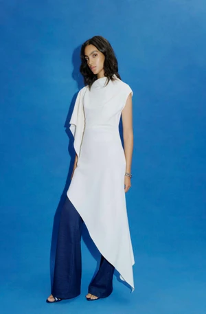 Model Noemi Kassa wearing a white asymmetrical drape tunic and wide-leg dark pants against a vibrant blue backdrop.