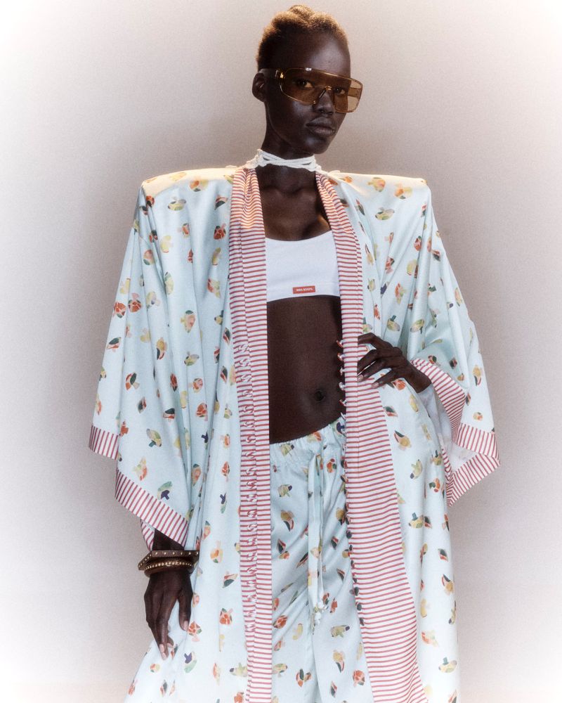 Model wearing an oversized printed kimono robe with shoulder pads, tinted sunglasses, and white crop top against a studio background.