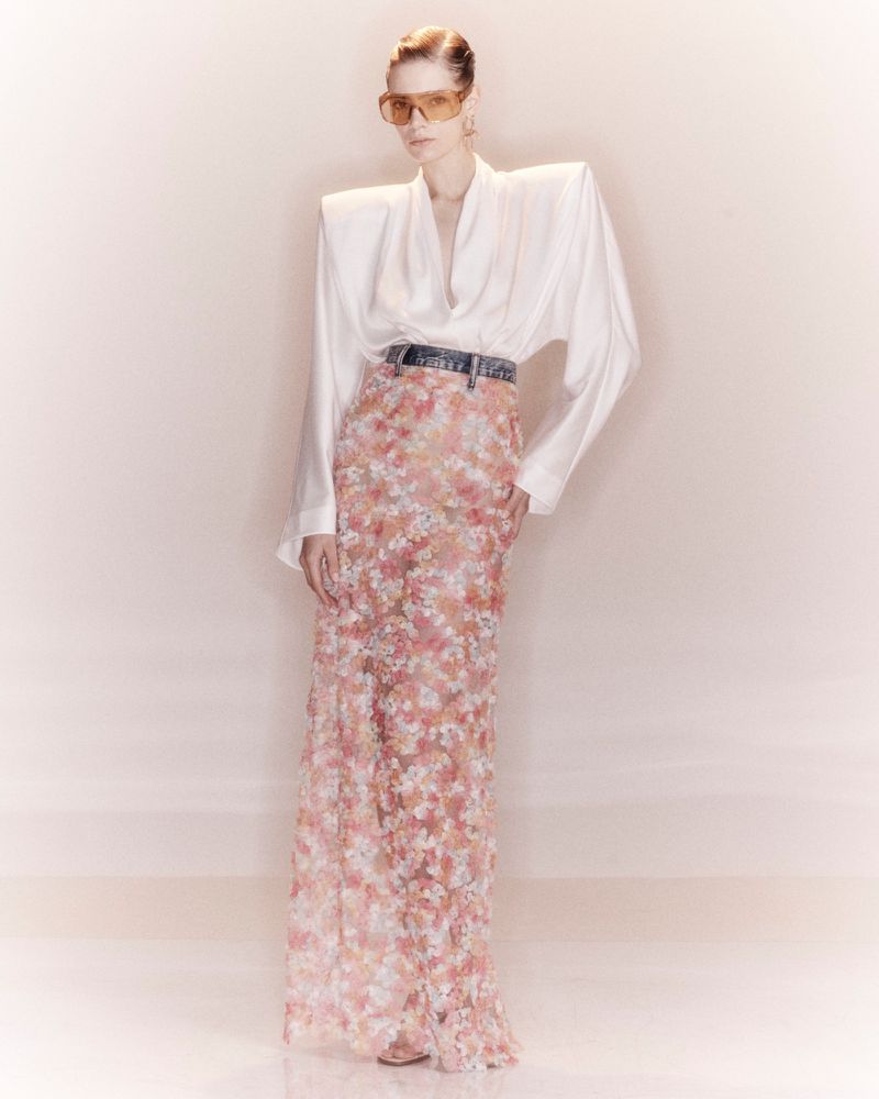 Model wearing a white structured shoulder blouse and a long floral skirt with tinted sunglasses against a pale studio backdrop.