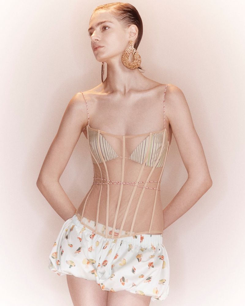 Model wearing a sheer corset top with visible boning and white printed bubble shorts against a warm neutral studio backdrop.