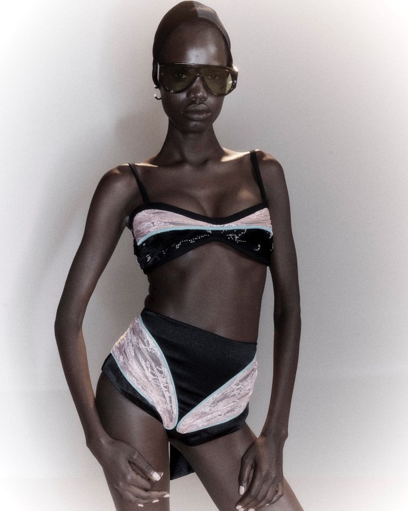 Model wearing dark shield sunglasses and a black high-waisted lace bikini set against a neutral studio backdrop.