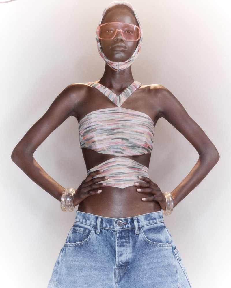 Model posing with hands on hips wearing a patterned cutout halter top, matching headpiece, tinted visor, and denim against a white studio background.