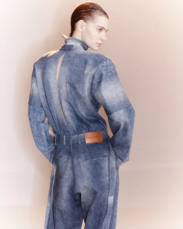 Model seen from behind wearing a high-neck faded blue denim jumpsuit with an open back detail against a neutral studio backdrop.