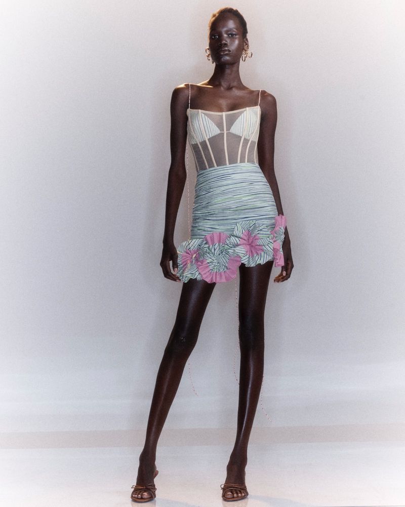Black female model in a sheer corset top and ruched mini skirt with pink ruffled accents posing on a white backdrop.