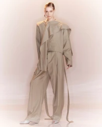 Model wearing an oversized draped beige suit with wide-leg trousers against a soft warm studio backdrop.